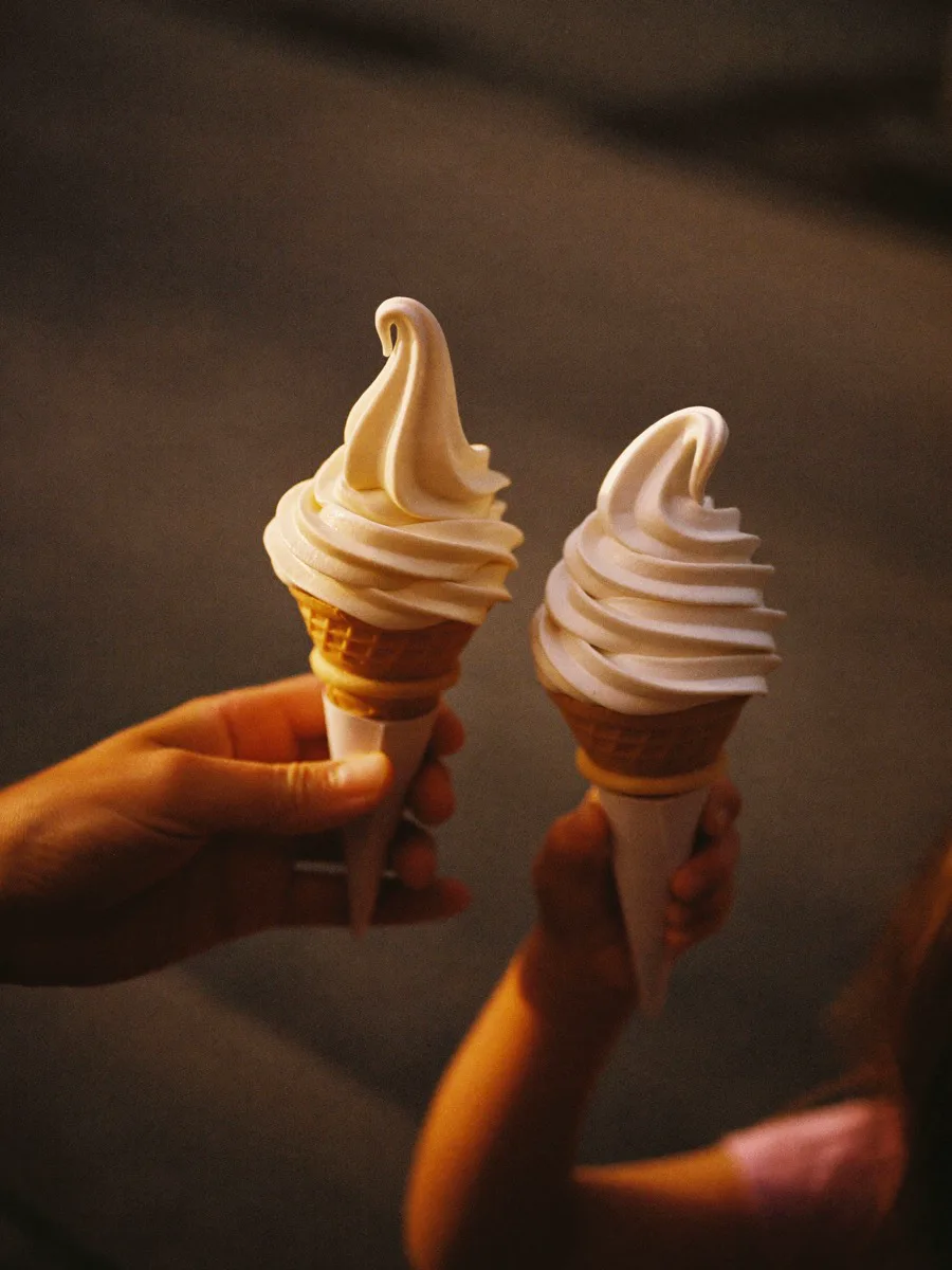 Two ice cream cones held up in warm evening light outside a small ice cream shop
