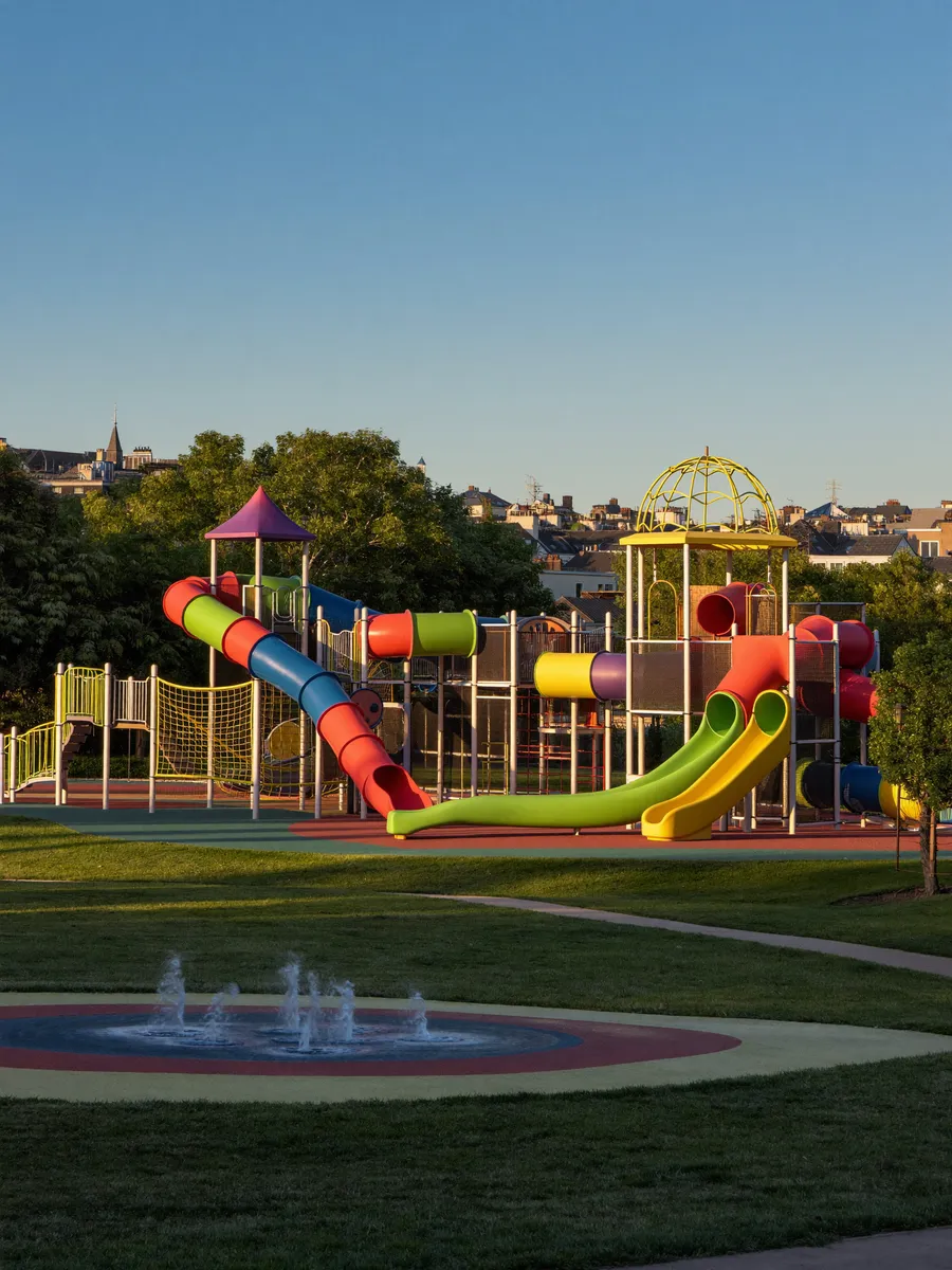 A sunlit neighborhood playground with swings and a slide among green trees